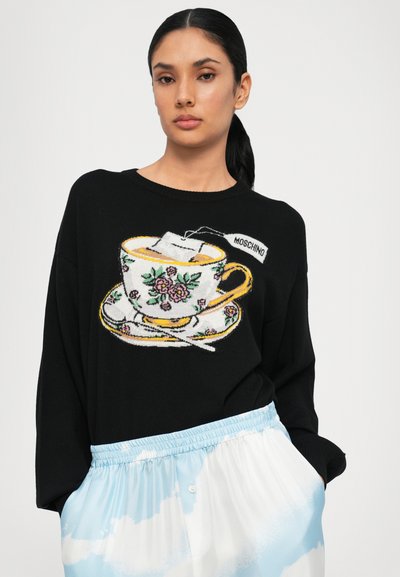Schwarzer Pullover mit einem farbenfrohen Design von einer Teetasse und einer Untertasse, verziert mit Blumenmustern, kombiniert mit blau-weißen Hosen mit Wolkenmuster.