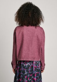 Burgundijska jeans jakna, skrajšana zasnova, z dolgimi rokavi in gumbi na manšetah, nosena čez cvetličasto obleko temnih tonov in živih barv.