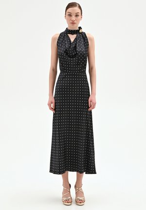 Donna con abito midi nero senza maniche a pois bianchi, scollo drappeggiato e dettaglio con fibbia dorata, indossa tacchi dorati con cinturini.
