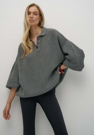 Blondynka nosi oversize szary sweter z dużymi guzikami i bufiastymi rękawami, zestawiony z czarnymi dopasowanymi spodniami, pozując na tle białej ściany.