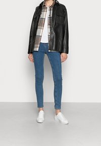 Svart läderjacka över en brun och vit rutiga skjorta, tillsammans med blå slim fit jeans och vita sneakers. Slät yta, avslappnad design.