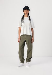 T-shirt blanche oversize avec un col rond, associée à un pantalon ample couleur olive. Des baskets blanches finalisent la tenue.