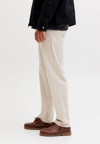 Bărbat purtând pantaloni bej, jachetă închisă la culoare și pantofi casual maro cu șireturi, stând în picioare lateral pe un fundal simplu și deschis la culoare.