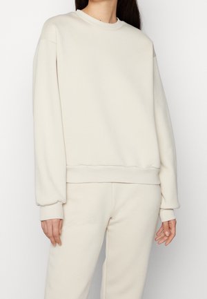 Personne portant un sweat-shirt à manches longues crème à col rond et un pantalon assorti, debout devant un fond uni.