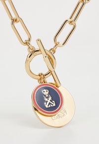 Zlatý tónovaný náhrdelník s velkými články ve tvaru svorky, dvěmi přívěsky: jeden oválný, zlatý s modrými a červenými akcenty, embosovaným logem a plochým diskem.