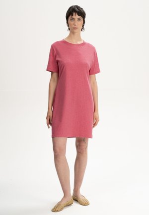 Vrouw met kort donker haar draagt een effen roze jurk met korte mouwen tot op de knie en beige gevlochten platte schoenen, staand tegen een witte achtergrond.