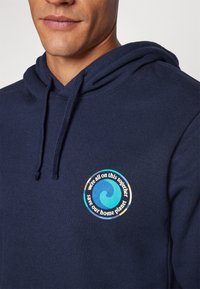 Hanorac din bumbac de culoare albastru marin, cu un patch grafic circular care prezintă un design cu valuri și text, subliniind prietenia față de mediu. Glugă ajustabilă cu șnur.