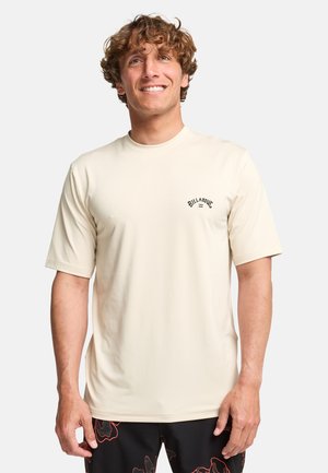 Uomo sorridente con capelli ricci che indossa una maglietta beige a maniche corte semplice e pantaloncini neri con motivi astratti rossi, in piedi davanti a uno sfondo bianco.