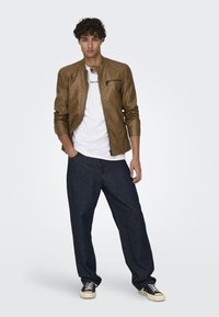 Giacca di pelle marrone con colletto rialzato, t-shirt bianca sotto e jeans di denim scuro larghi. Sneakers bianco e nere completano il look.