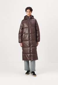 Abrigo acolchado metálico marrón con capucha, con un diseño hasta la rodilla, textura acolchada, cremallera frontal y bolsillos laterales. Lleva puesto unos jeans azules.