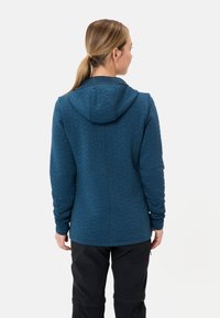 Blauwe gewatteerde hoodie met een capuchon, voorzien van een textuurpatroon, lange mouwen en een aangesloten design. Draagt in combinatie met donkere broek.