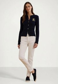 Mulher em pé com os braços relaxados, vestindo um cardigan azul-marinho com botões e um logotipo dourado, calças crème e sapatilhas pretas, em um fundo simples.