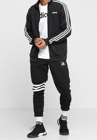 Black Adidas tracksuit with white stripes on sleeves and pants; featuring a zip-up jacket, white t-shirt, and black athletic shoes.