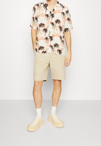 Camicetta a maniche corte di colore chiaro con stampa di palme; pantaloni corti beige; calze bianche; scarpe platform beige. Outfit estivo casual.