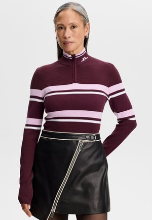 Woman with long gray hair wearing a maroon and pink striped pullover with zipper and a black asymmetrical leather skirt.