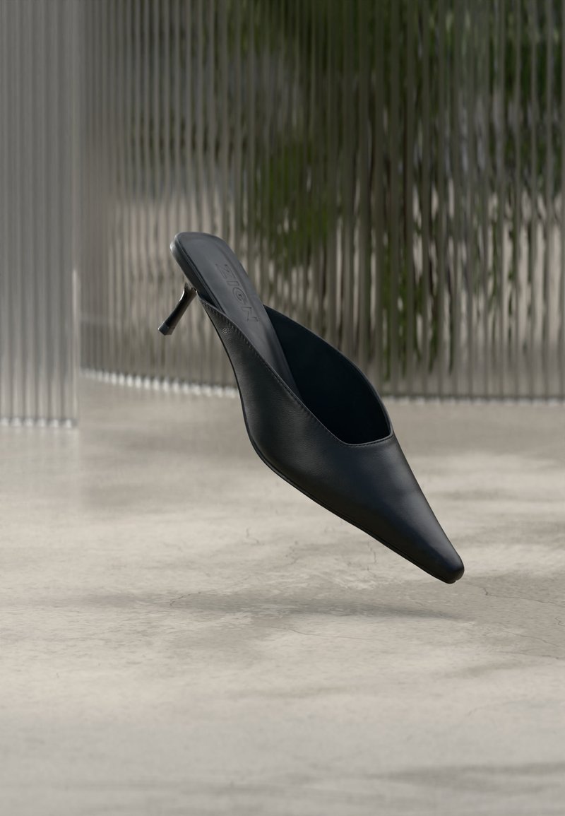 Mule noire à bout pointu avec un talon fin et haut flottant au-dessus d'un sol en béton lisse, avec un arrière-plan flou à rayures verticales.