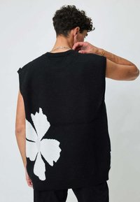 Pull en tricot sans manches noir avec un grand motif floral blanc au dos. Le design est simple et audacieux, avec une finition texturée.