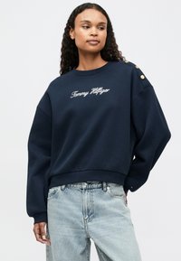 Marineblå hettegenser med brodert "Tommy Hilfiger"-logo og gullknappdetaljer på skuldrene; avslappet passform og ribbestrikkede mansjetter.