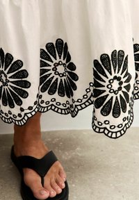 Witte rok met zwarte bloemenborduursels aan de zoom. Voorzien van geschulpte randen en fijne stiksels. Zwarte teensandalen zichtbaar.