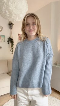 Suéter de malha azul clara com textura canelada nas mangas e na bainha. Apresenta ombros caídos e um decote redondo. Usado com calças brancas.