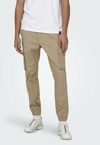 Pantalons cargo beiges avec poignets élastiques, plusieurs poches latérales et une coupe décontractée. Portés avec un t-shirt blanc et des baskets blanches.