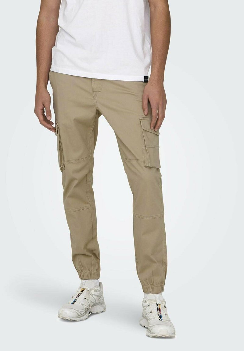 Pantalons cargo beiges avec poignets élastiques, plusieurs poches latérales et une coupe décontractée. Portés avec un t-shirt blanc et des baskets blanches.