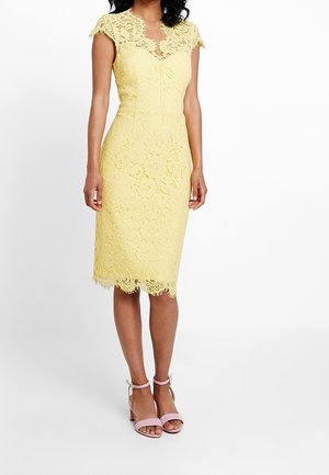 Frau trägt ein gelbes Spitzenkleid bis zum Knie mit kurzen Ärmeln und rosa Knöchelriemen-Absätzen, steht vor einem schlichten weißen Hintergrund.