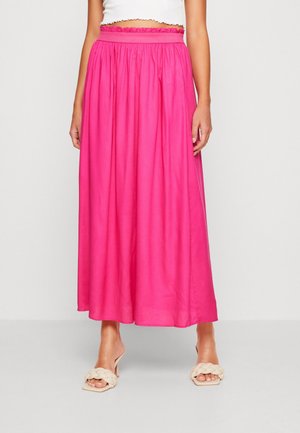 Kvinde iført en stærkt pink, højtaljet, ankel-lang nederdel med en hvid crop top og beige flettede sandaletter med hæl.