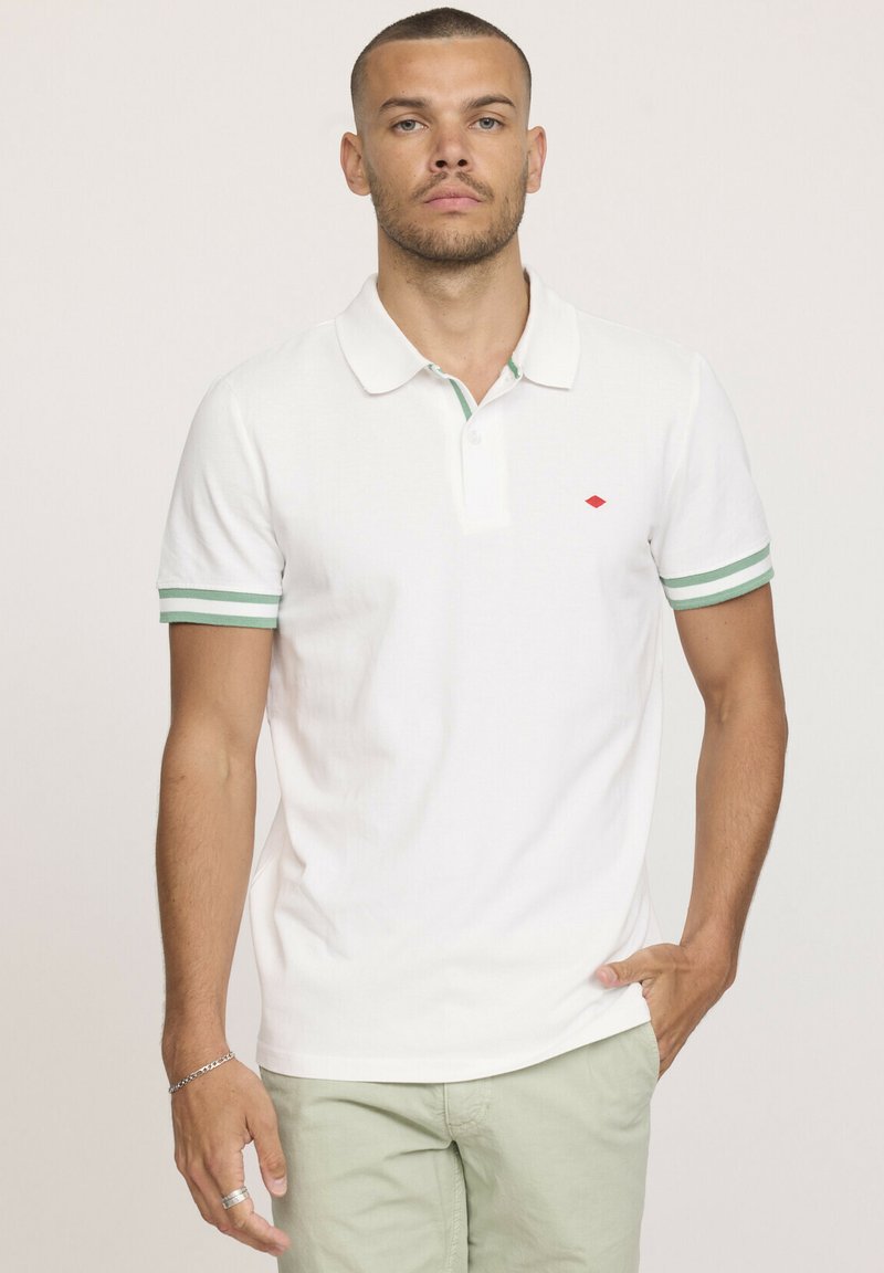 Chemise polo blanche avec des poignets et un col à rayures vertes et blanches, et un petit logo en losange rouge sur la poitrine gauche. Manches courtes.
