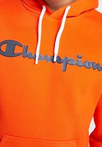 Sudadera naranja de tela suave, con un gran logo de "Champion" en color marino. Tiene cordones blancos y un bolsillo tipo canguro.
