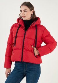 Rød pufferjakke med hætte, sort lynlås og snoretræk. Har sidelommer og et quiltet mønster. Bæres over en sort turtleneck.