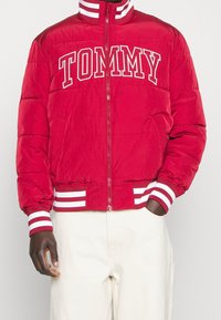 Röd puffjacka med hög krage, frontdragkedja, vit "TOMMY" logo och randiga ribbade ärmslut och midja. Mjuk textur, isolerat design.