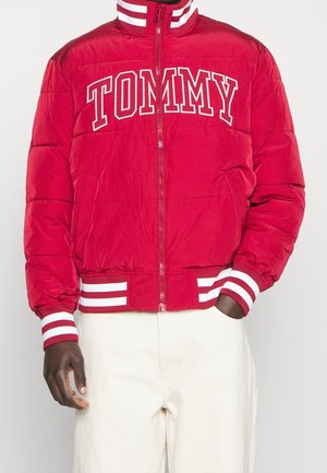 Veste rembourrée rouge avec un col haut, fermeture éclair frontale, logo "TOMMY" blanc, et poignets et ceinture côtelés rayés. Texture douce, design isolé.