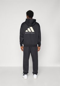 Schwarze Kapuzenjacke mit großem beigem Adidas-Logo und dem Text "BASKETBALL" auf dem Rücken, kombiniert mit passenden schwarzen Jogginghosen. Weiße Sportschuhe.