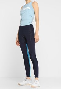 Hellblaues Sporttanktop mit weißem Logo, kombiniert mit marineblauen Leggings mit türkisem Streifen und strukturiertem Muster. Weiße Sneakers.