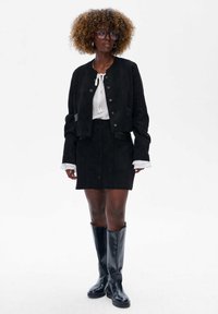 Zwarte suède cropped jas met geribbelde details, gecombineerd met een zwarte suède knopenrok. Zwarte kniehoogte laarzen maken de outfit compleet.