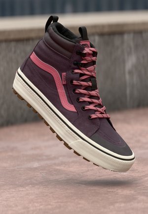 Lila und pinke Vans High-Top Sneaker mit gemusterten Schnürsenkeln, die über einer Betonoberfläche im Freien schweben.