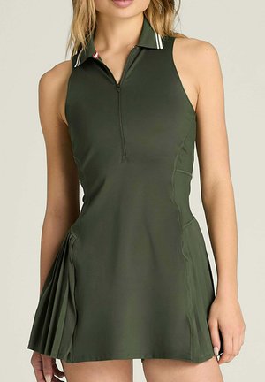 Mujer vistiendo un vestido de tenis sin mangas de color verde oscuro con cuello con cierre y detalle plisado en un lado.