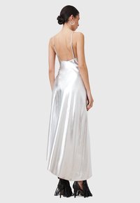 Abito maxi in satin metallico argento con sottili spalline regolabili, schiena scoperta e una silhouette fluida. Abbinato a scarpe con tacco a frange nere.