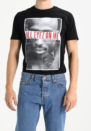 Mann trägt ein schwarzes T-Shirt mit einem schwarz-weißen Gesichtsfoto und rotem Text "ALL EYEZ ON ME", kombiniert mit blauer Jeans.