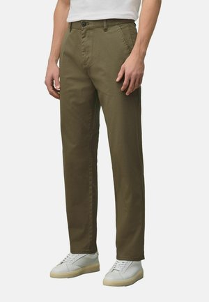 Mann trägt olivgrüne gerade geschnittene Hose und weiße Turnschuhe, steht vor einem einfarbigen Hintergrund.