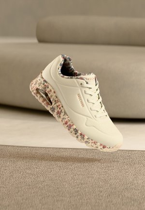 Baskets Skechers couleur crème avec semelle et doublure intérieure à motif floral, flottant au-dessus d'un tapis beige texturé dans une pièce aux tons neutres.