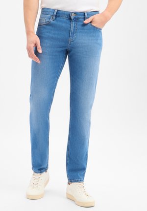 Mann trägt hellblaue Slim-Fit-Jeans mit fünf Taschen und weißen Sneakers, steht mit einer Hand in der Tasche vor weißem Hintergrund.
