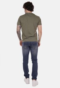 Camicia verde oliva a maniche corte con una vestibilità rilassata, abbinata a jeans a gamba dritta blu scuro e sneakers bianche. Tatuaggi visibili sulle braccia.