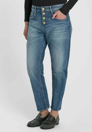 Midrise blaue Denim-Jeans mit vier goldenen Knöpfen, getragen mit schwarzen Loafers und einem schwarzen Langarmtop, Hand in der Tasche.
