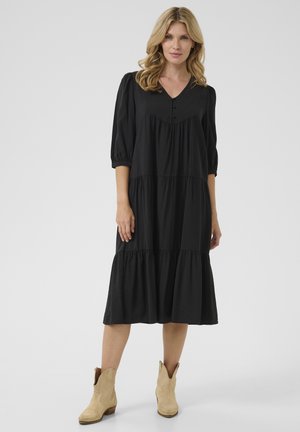 Schwarzes schichtetes Kleid mit dreiviertel Puffärmlen, V-Ausschnitt und Knopfdetails. Weicher Stoff mit fließender Silhouette, kombiniert mit beigen Stiefeln.