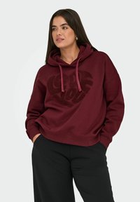 Maroon hooded sweatshirt dengan desain hati hitam bertekstur. Dilengkapi dengan tali serut yang dapat disesuaikan dan manset bergaris, dipadukan dengan celana hitam.