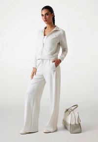 Hvid lynlåstrøje og matchende bukser med striber, lavet af blødt materiale. Fuldendes af en lysegrå håndtaske med lynlås.