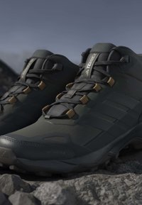Μαύρα και σκουρογκρι Adidas Terrex πεζοπορικά παπούτσια με καφέ θηλιές κορδονιών τοποθετημένα σε βραχώδες έδαφος σε εξωτερικό χώρο.