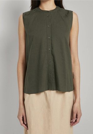Personne portant un haut sans manches kaki boutonné et une jupe beige clair, debout devant un fond uni clair.